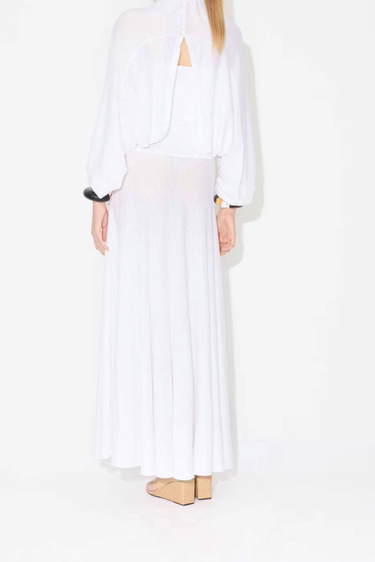 Back view of white linen maxi dress on model wearing white pullover and natural wedge sandals, against white background.