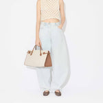 Front view of light blue denim pants with model wearing knotted white top and square ballet flats with brown and creme handbag on white background.