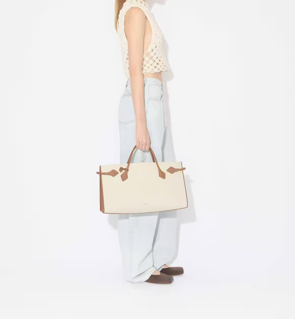 Side view of light blue denim pants on model wearing knotted white top with brown square ballet flats and creme/brown handbag on white background.