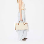 Side view of light blue denim pants on model wearing knotted white top with brown square ballet flats and creme/brown handbag on white background.