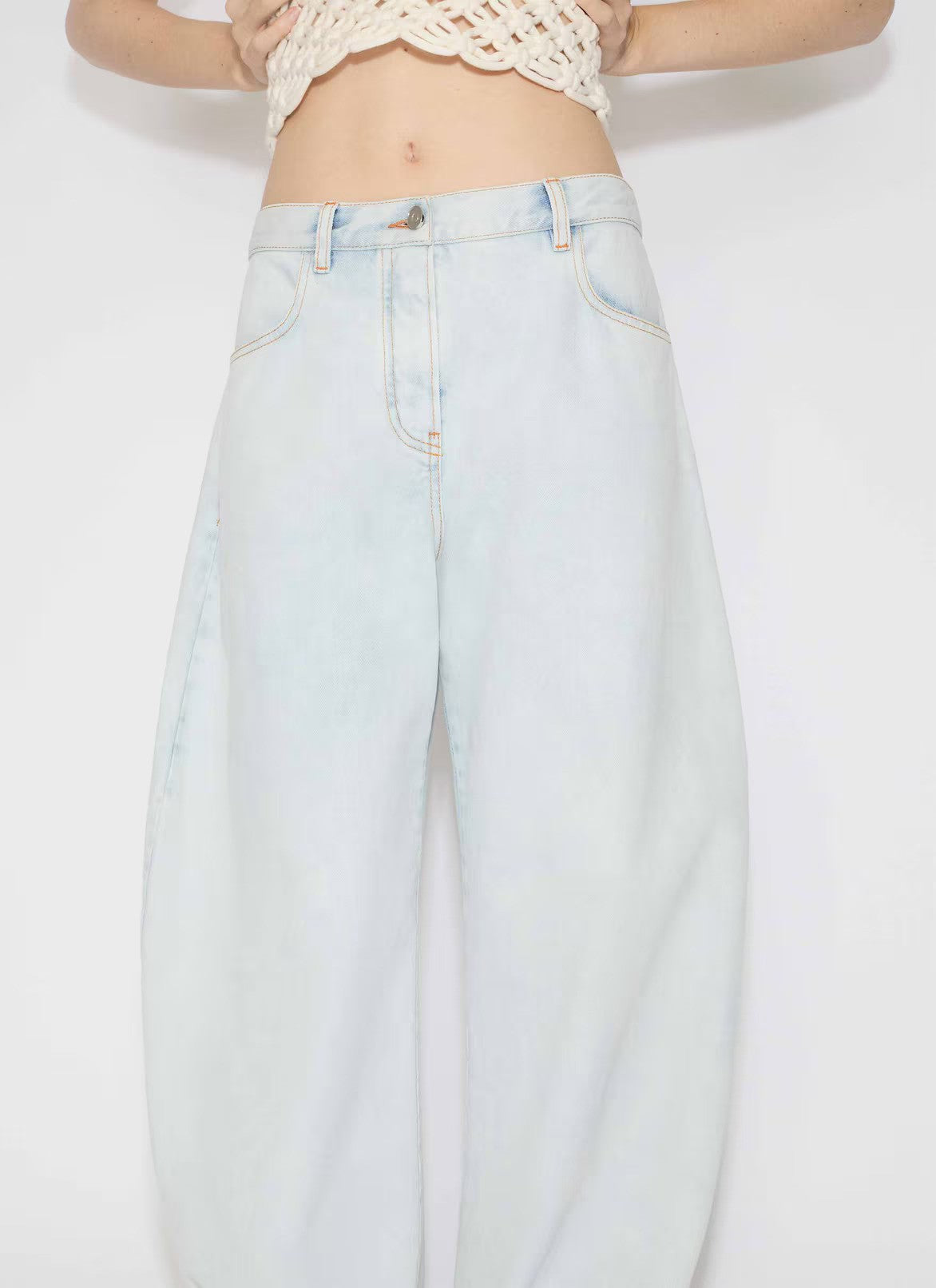 Front closeup view of light blue denim pants on model wearing knotted white top on white background.