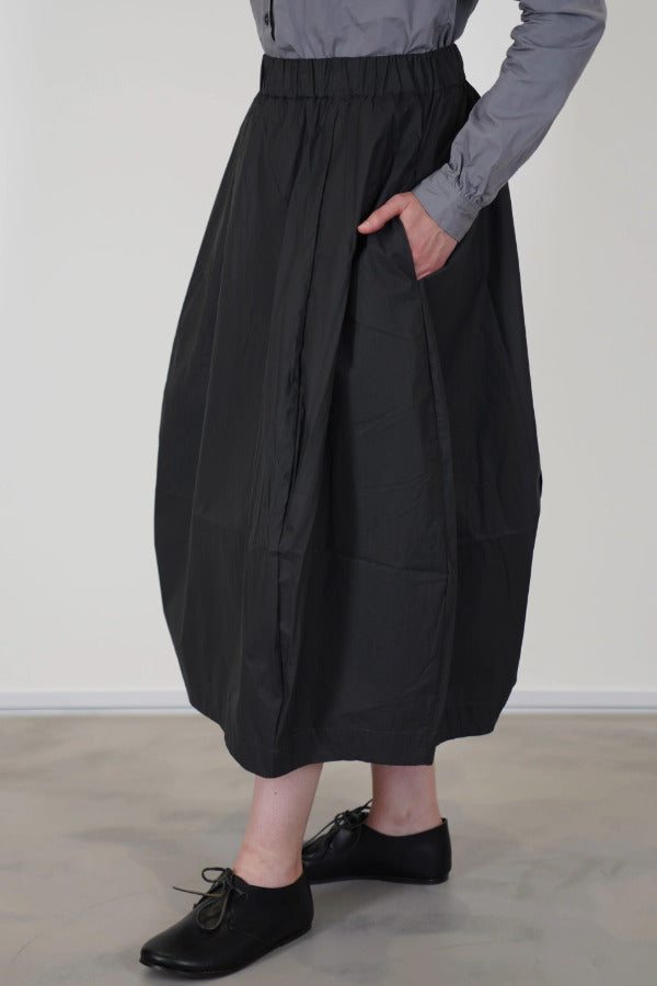 Side view of carbon carla skirt on model wearing dark grey top and black oxfords, against a white background.