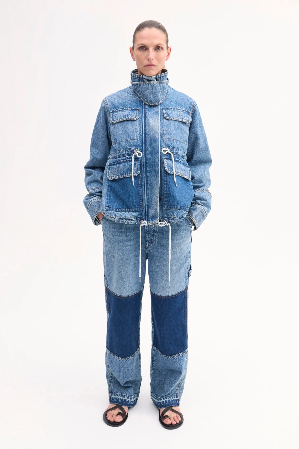 Front view of cropped military jacket on model wearing patchwork denim pants and black sandals on white background.