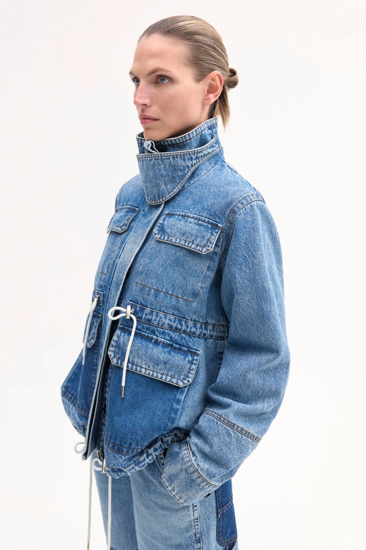 Side Closeup view of cropped military jacket on model wearing denim patchwork pants on white background.