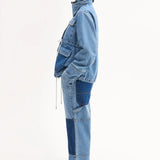 Side view of cropped military jacket on model wearing denim patchwork pants and black sandals on white background.