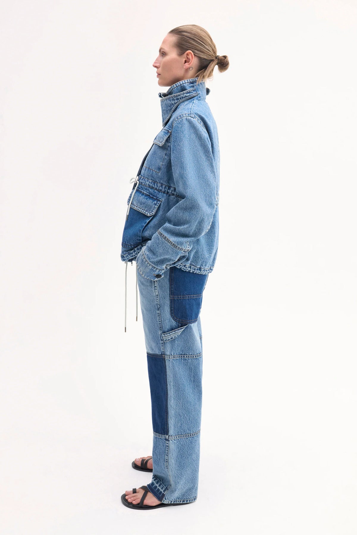 Side view of cropped military jacket on model wearing denim patchwork pants and black sandals on white background.