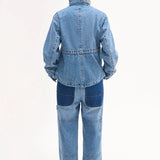 Back view of cropped military jacket on model wearing denim patchwork pants on white background.