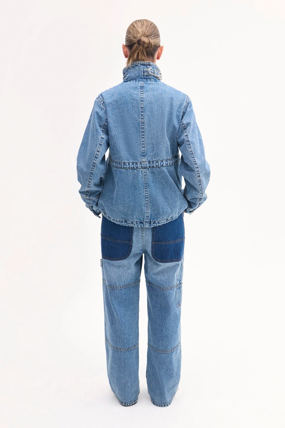 Back view of cropped military jacket on model wearing denim patchwork pants on white background.