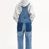 Back view of indigo overalls on model wearing grey hoodie and black sandals on white background.