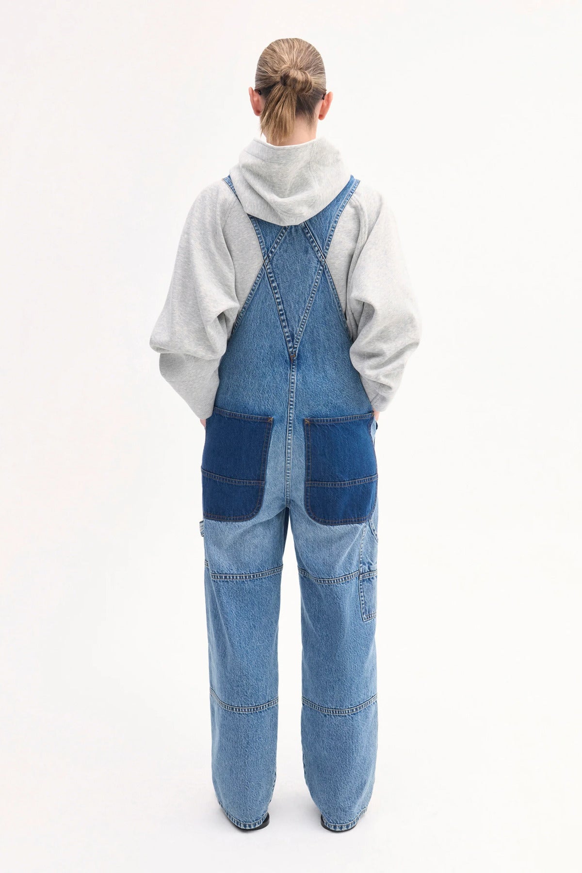 Back view of indigo overalls on model wearing grey hoodie and black sandals on white background.
