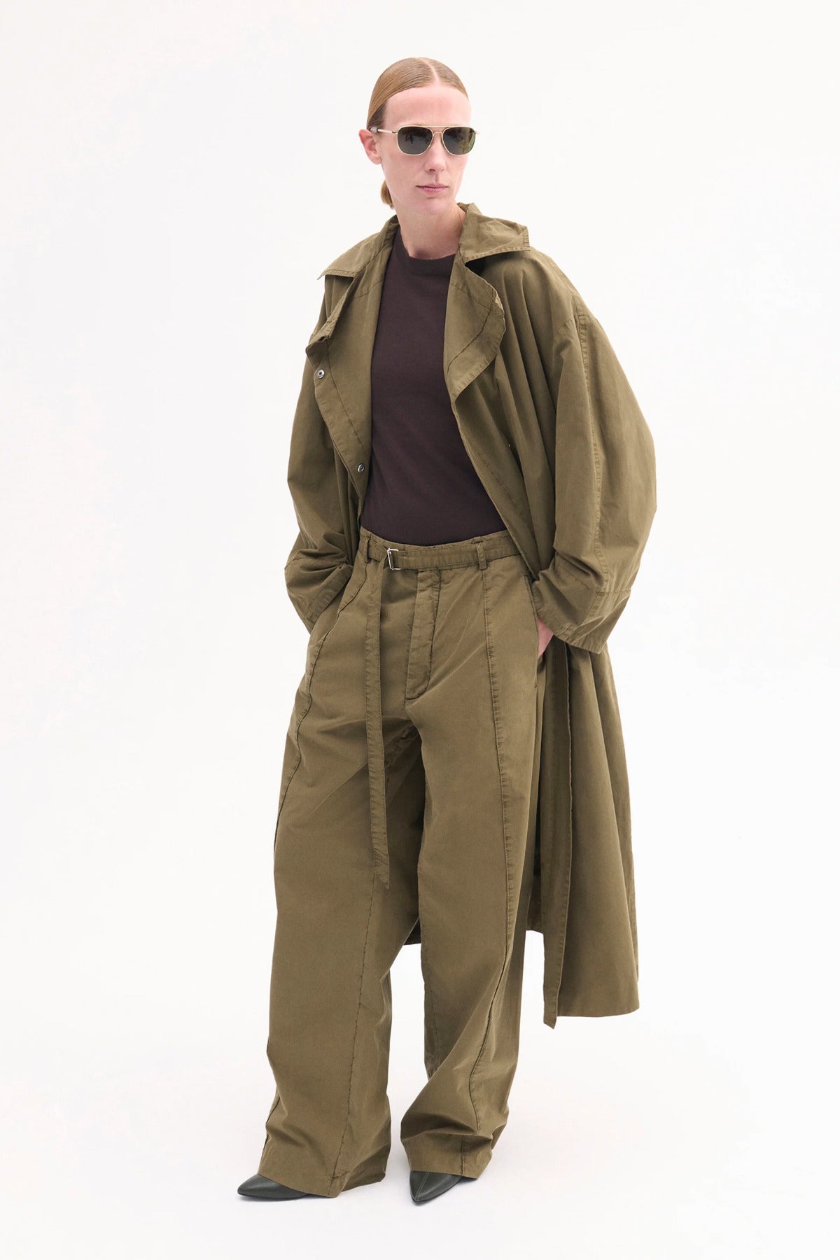 Front view of olive oversized trench coat on model wearing brown shirt, olive trousers and black pointed toe heels on white background.