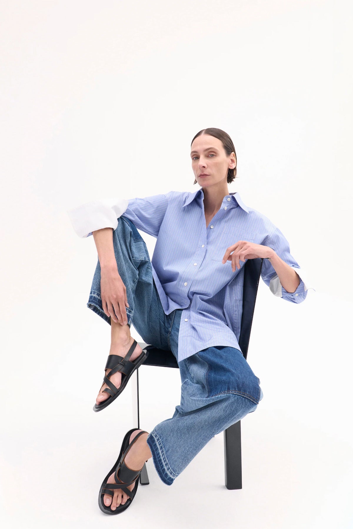 A model with light brown hair is shown in the black leather shoes sitting in a chair grabbing her knee to show the shoe with a white background.