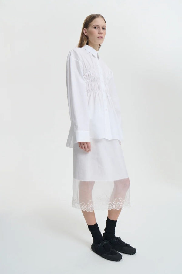 Side view of white braxton shirt on model wearing white slip and black sneakers, against a white background.