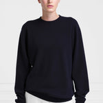 Front view of navy you sweater on light haired model wearing white pants on white background.