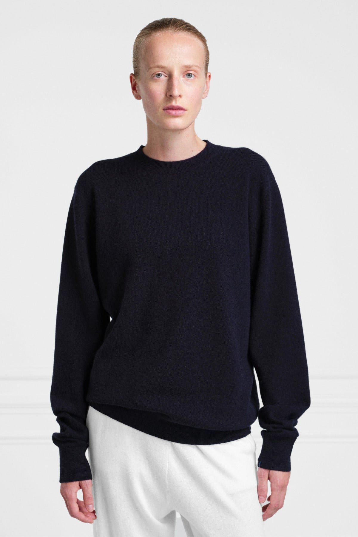 Front view of navy you sweater on light haired model wearing white pants on white background.