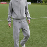 Front view of grey no 320 rush trouser on model wearing grey jacket and sneakers, against a grassy field background.