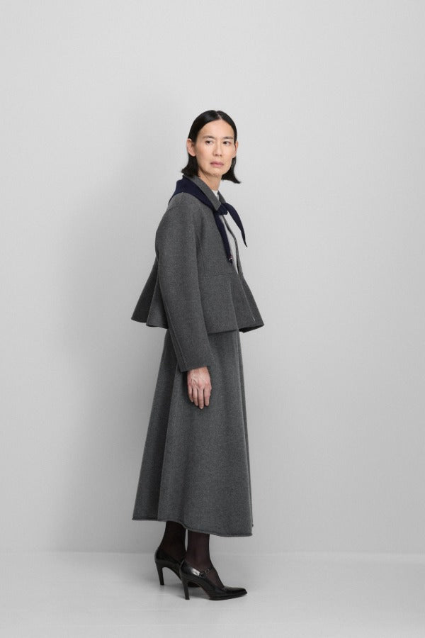 Side view of felt no 442 queen coat on model wearing grey skirt and heels, against a grey background.