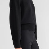 Side view of black lofty lara sweater on model wearing balck pants, against a white background.