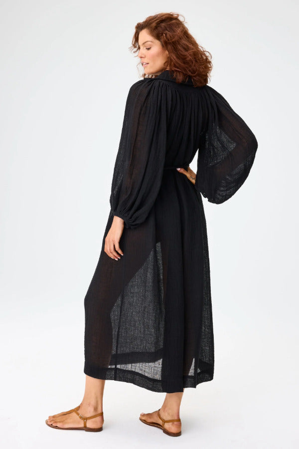 Slanted back view of black maxi poet dress on brown-haired model wearing brown sandals, against a white background.