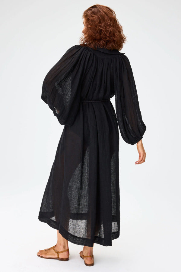 Back view of black maxi poet dress on brown-haired model wearing brown sandals against a white background.