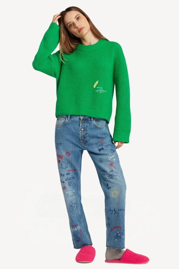 Front view of denim embroidered scribble jean on model wearing green sweater and pink slides, against a white background.