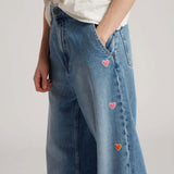 Side closeup view of mid blue embroidered straight leg jean on model wearing white t-shirt, against a white background.
