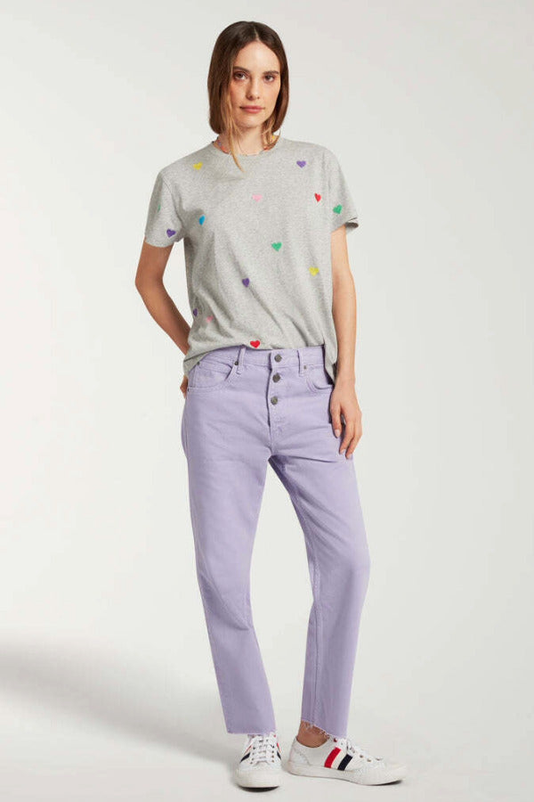 Front view of lilac garment dyed jeans on model wearing grey shirt with hearts and white sneakers, against white background.
