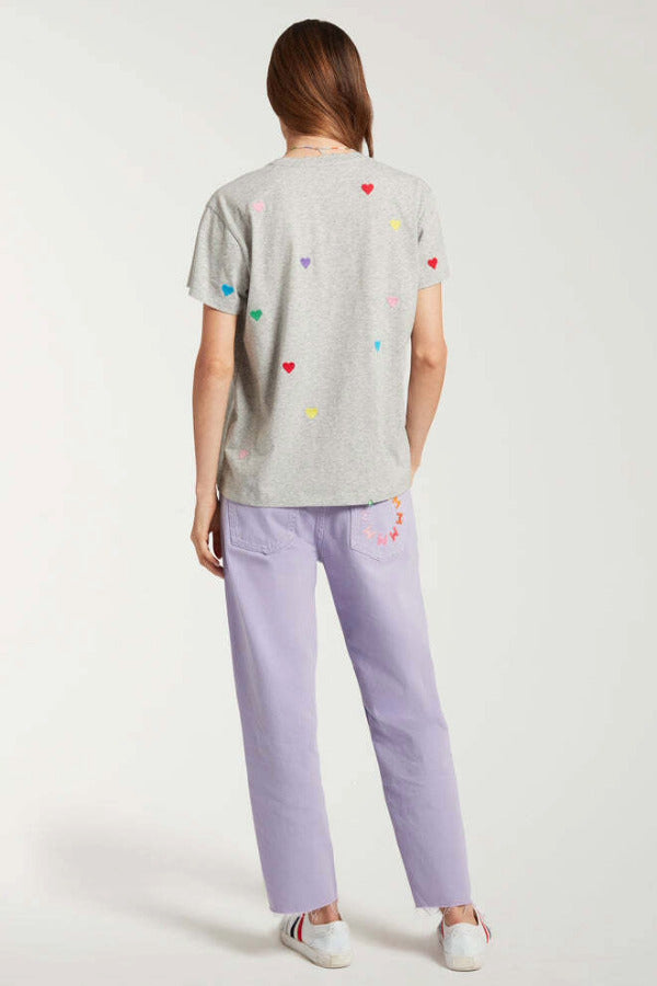 Back view of lilac garment dyed jeans on model wearing grey shirt with hearts and white sneakers, against a white background.