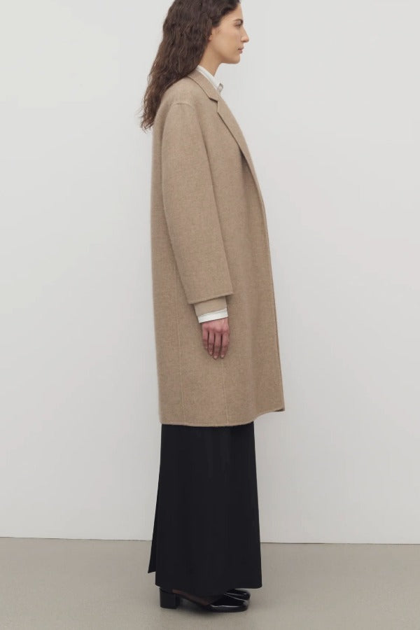 Side view of natural fedra coat on model wearing black skirt and mules, against a white background.