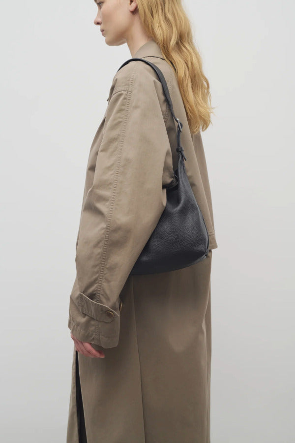 Back side of black isa shoulder bag on model wearing khaki trench coat, against white background.