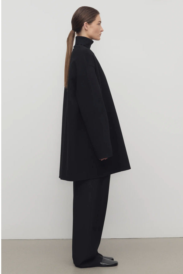 Side view of black kala coat on model wearing black turtleneck and trousers, against a white background.