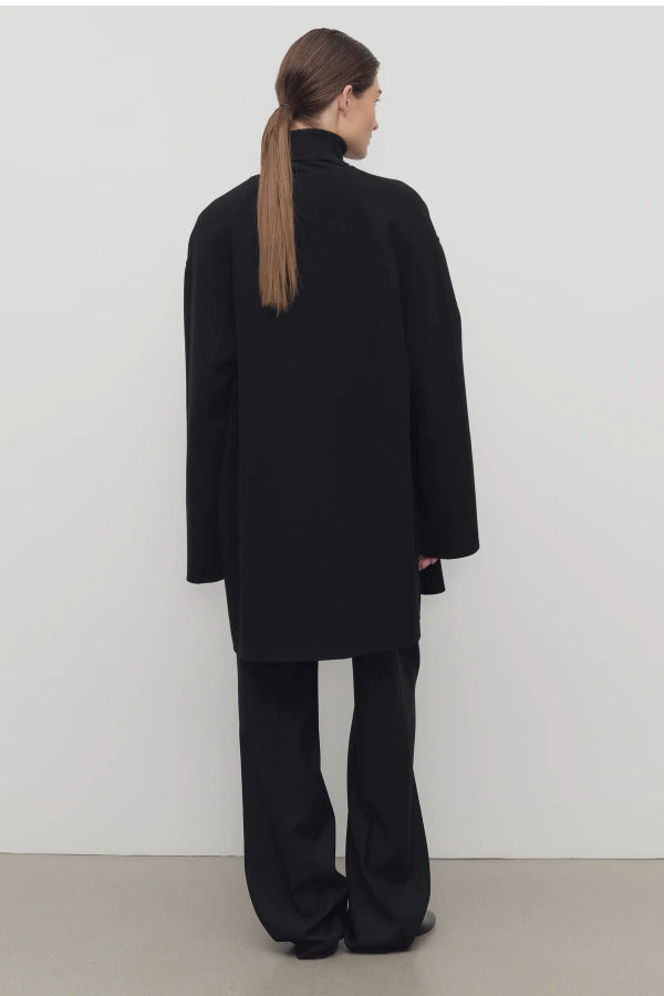Back view of black kala coat on model wearing black turtleneck and trousers, against a white background.