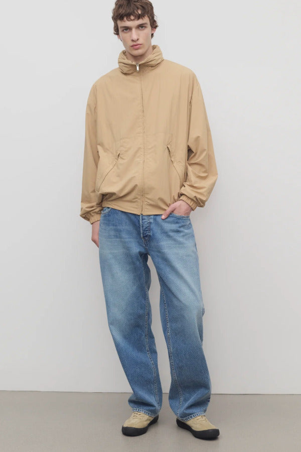 Front view of medium blue men's ross jean on model wearing sand windbreaker and sneakers, against a white background.