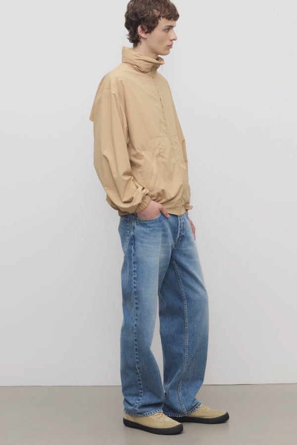 Side view of medium blue men's ross jean on model wearing sand windbreaker and sneakers, against a white background.