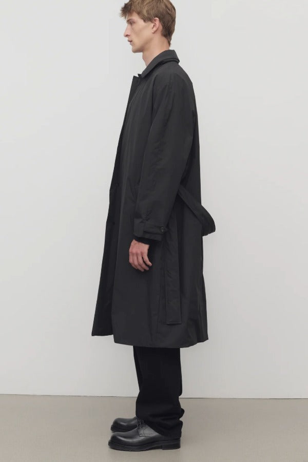 Side view of black tarian coat on model wearing black pants, shirt and boots, against a white background.