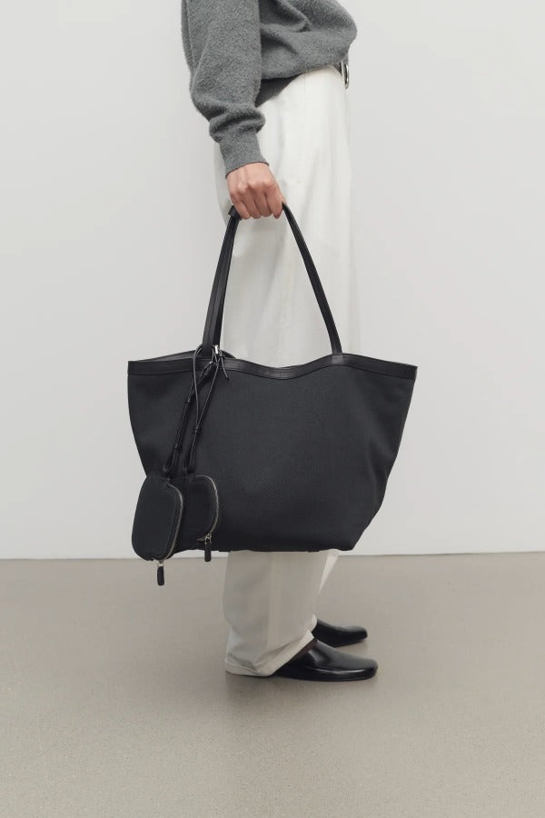 Front view of black park tote three canvas on model wearing white pants and grey sweater with mules, against a white background.
