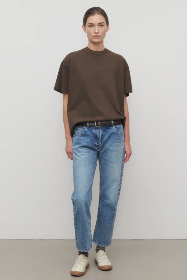 Front view of medium blue riaco jean on model wearing brown shirt and white sneakers, against a white background.