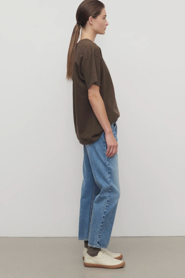 Side view of medium blue riaco jean on model wearing brown shirt and white sneakers, against a white background.