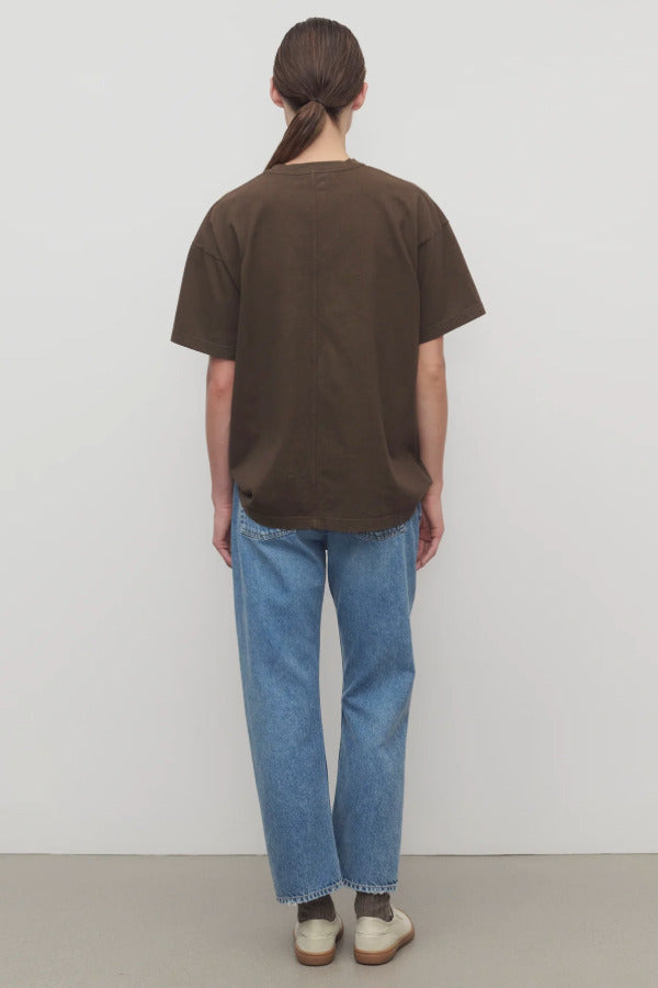 Back view of medium blue riaco jean on model wearing brown shirt and white sneakers, against a white background.