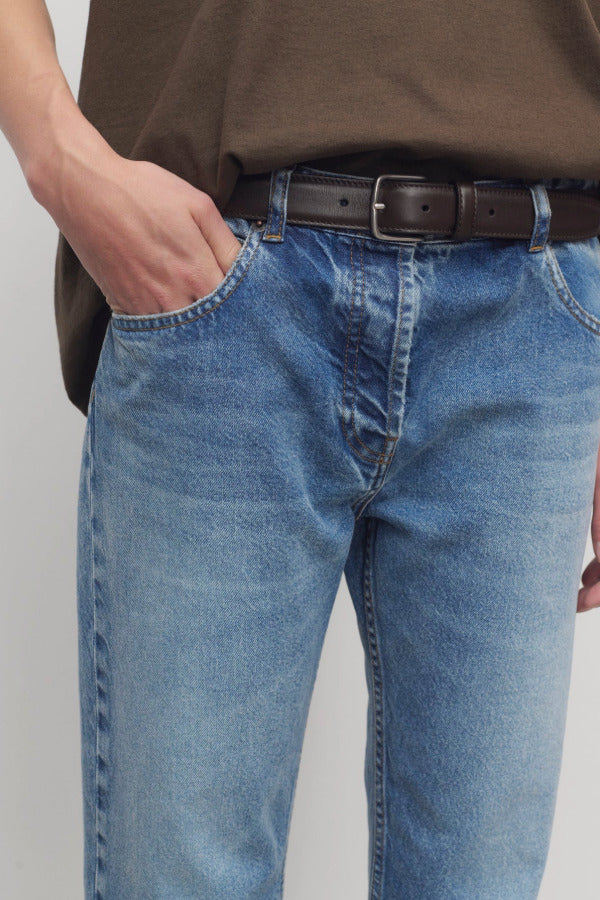Front closeup view of medium blue riaco jean on model wearing brown shirt, against a white background.