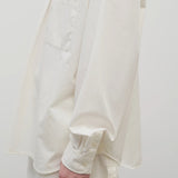 Side closeup view of white erika shirt on model wearing white trousers, against white background.