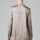 Back view of stone stretch silk satin regular fit shirt on model wearing chalk pants against a grey background