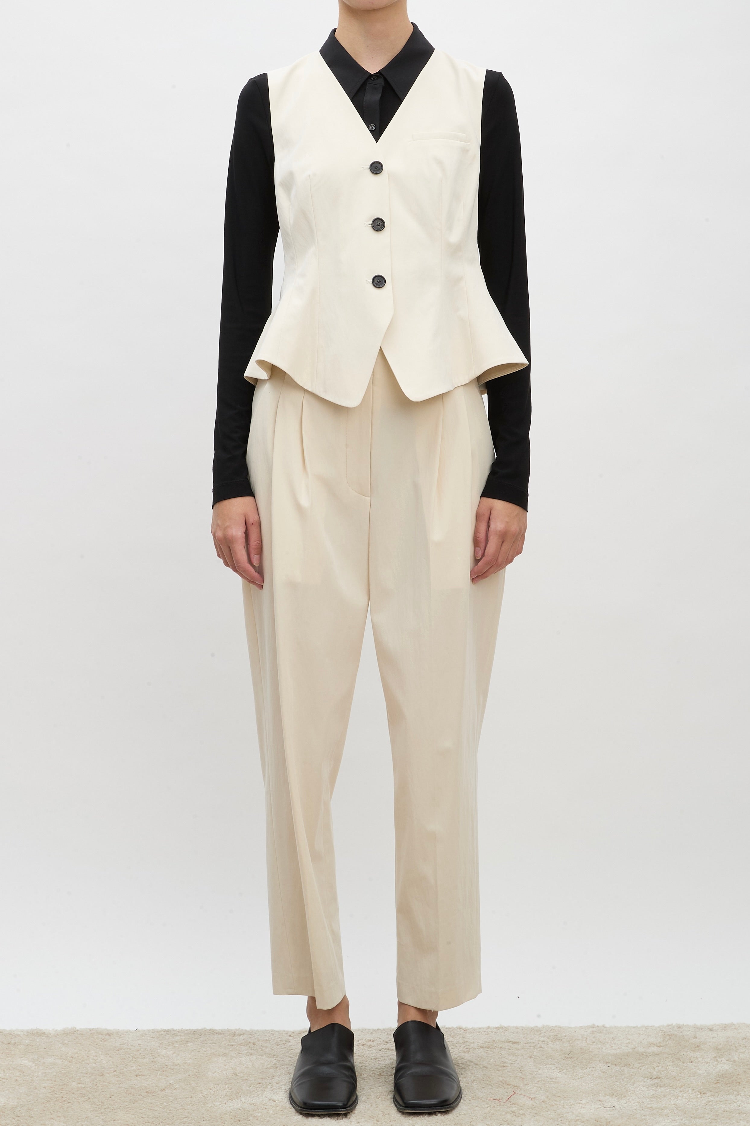 Front view of ivory raine pants on model wearing black shirt, ivory vest and brown loafers, against a white background.