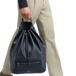 Front view of navy newport shoulder bag on model wearing khaki trousers, a navy sweater and brown sneakers, against a white background.
