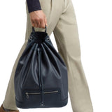 Front view of navy newport shoulder bag on model wearing khaki trousers, a navy sweater and brown sneakers, against a white background.