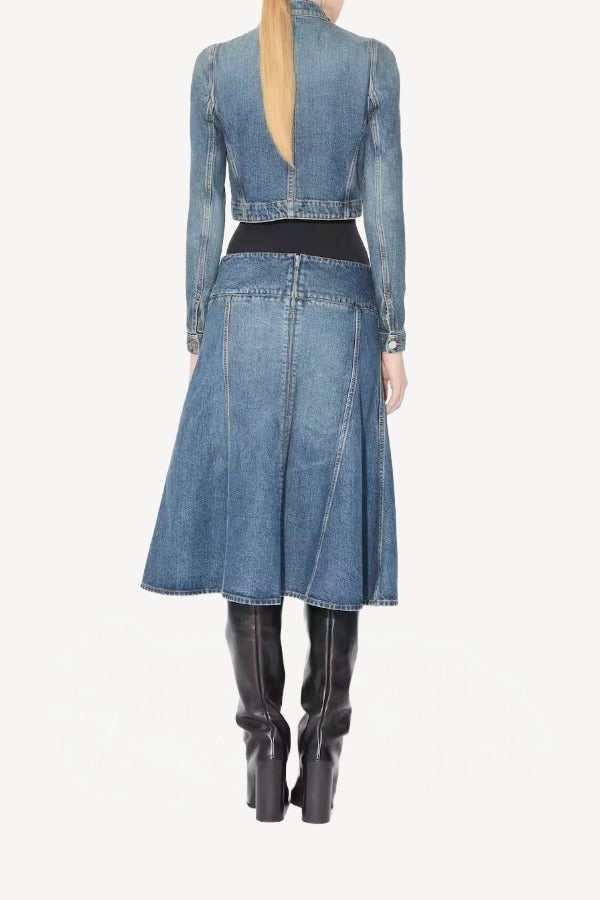 Back view of denim cropped jacket on model wearing black bodysuit, denim skirt, and boots, against a white background.