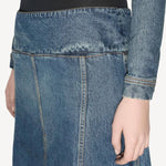 Side closeup of denim slit skirt on model wearing black bodysuit and denim jacket, against a white background.