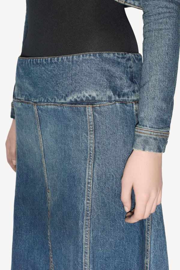 Side closeup of denim slit skirt on model wearing black bodysuit and denim jacket, against a white background.
