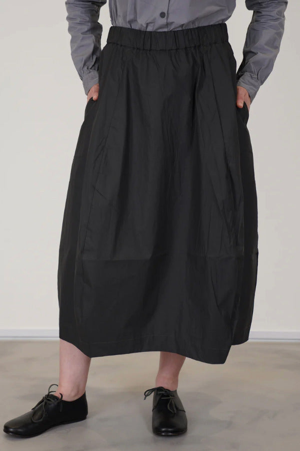 Front view of carbon carla skirt on model wearing dark grey top with black oxfords, against a white background.