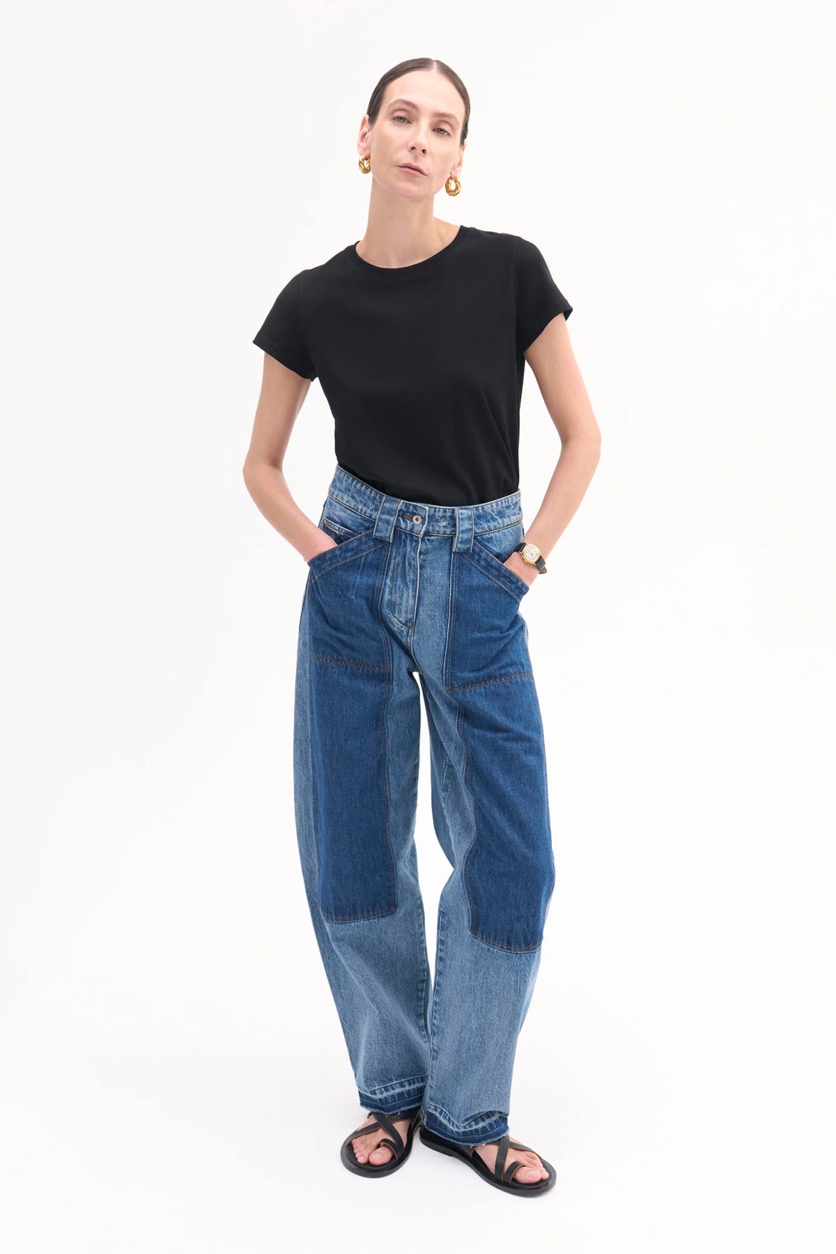 Front view of indigo denim patchwork pants on model wearing black t-shirt and black sandals, against white background.